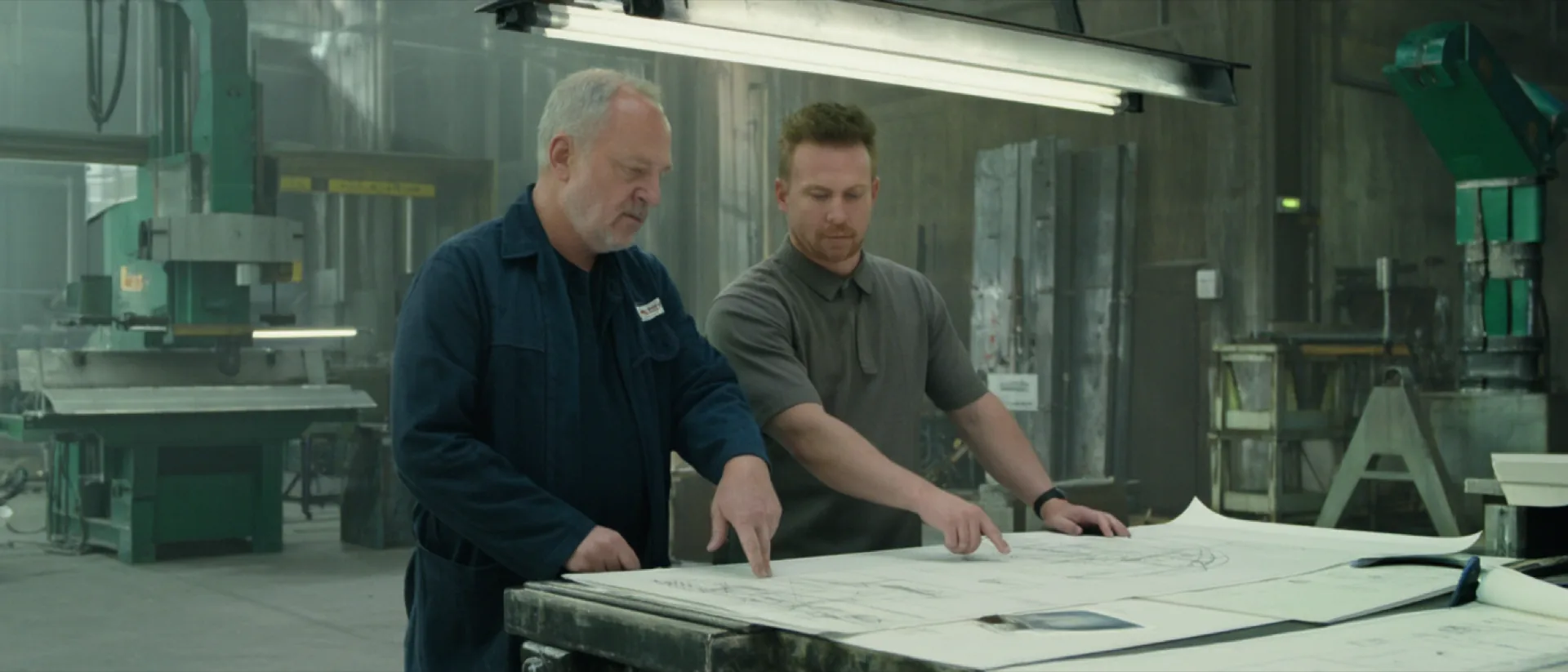 A shop owner and a consultant leaning over a paper production plan on a steel workbench in a small metal fabrication workshop